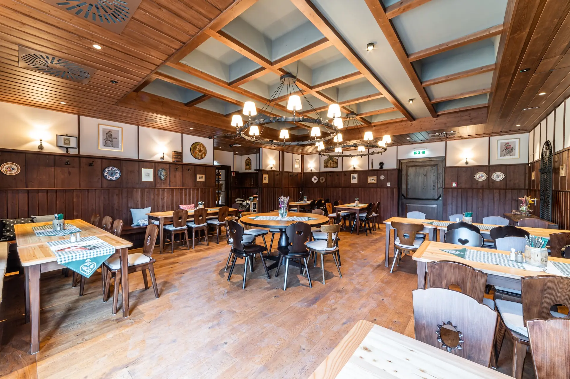Ein geräumiger, rustikaler Speisesaal mit Holzböden, -wänden und -decken, runden und rechteckigen Tischen, Holzstühlen, Kronleuchtern und traditionellem Dekor an den Wänden. Der Raum ist gut beleuchtet und übersichtlich gestaltet.