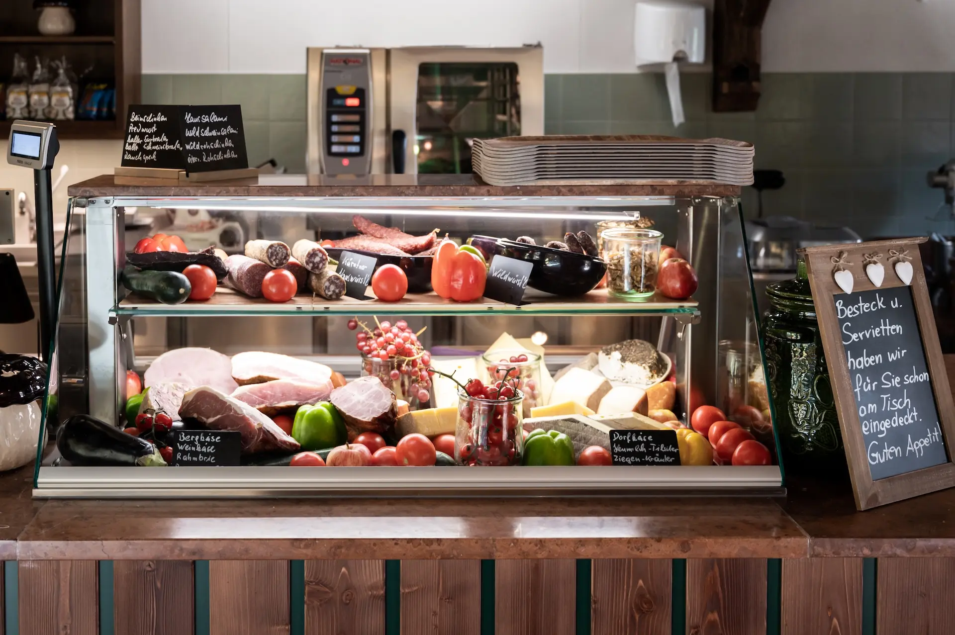 Eine Feinkosttheke bietet verschiedene Fleisch-, Käse- und Wurstsorten, Gemüse und Gläser an. Kreidetafeln mit Speisekarten und eine Holztheke sind in dem gemütlichen, rustikalen Ladeninneren zu sehen.