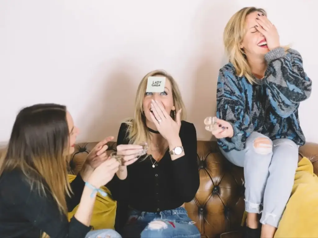 Drei lachende Frauen auf einer Couch, die Partyspiele für Erwachsene spielen - eine mit einem Lady-Gaga-Klebezettel auf der Stirn, eine andere, die ihren Mund bedeckt, und die dritte, die ihr Gesicht versteckt, während sie ein Schnapsglas hält.