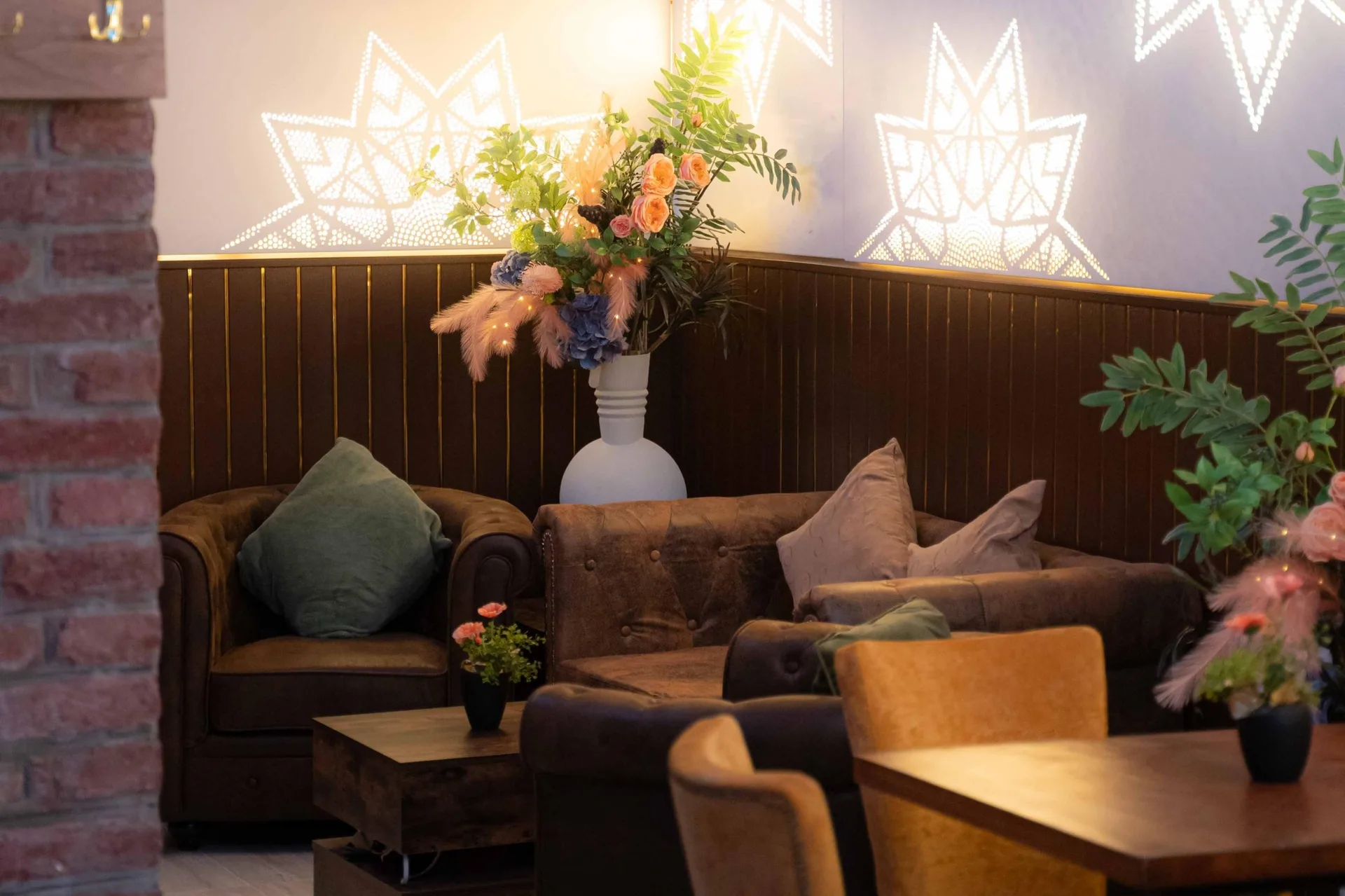 Eine gemütliche Café-Ecke mit braunen Sesseln, einem Holztisch, Zierpflanzen und einem großen Blumenarrangement in einer weißen Vase. Sanfte Stimmungsbeleuchtung in Kombination mit geometrischen Wandmustern schafft eine warme, einladende Atmosphäre.