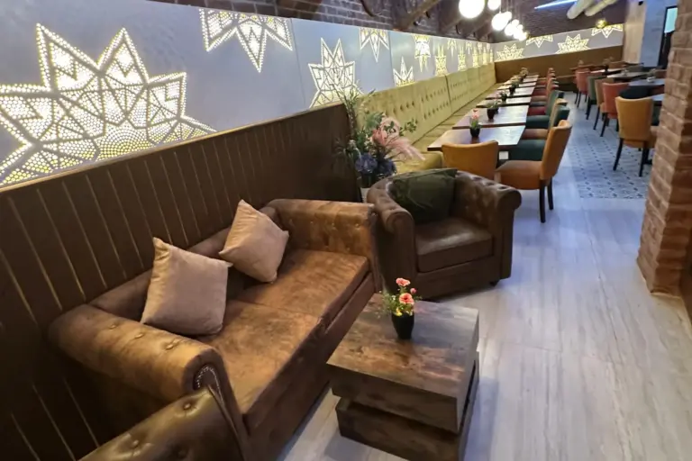 Eine gemütliche Lounge mit braunen Ledersofas, einem Couchtisch aus Holz, Reihen bunter Stühle und Tische, dekorativen Sternmustern an einer hinterleuchteten Wand und Pflanzen, die zur einladenden Atmosphäre beitragen.