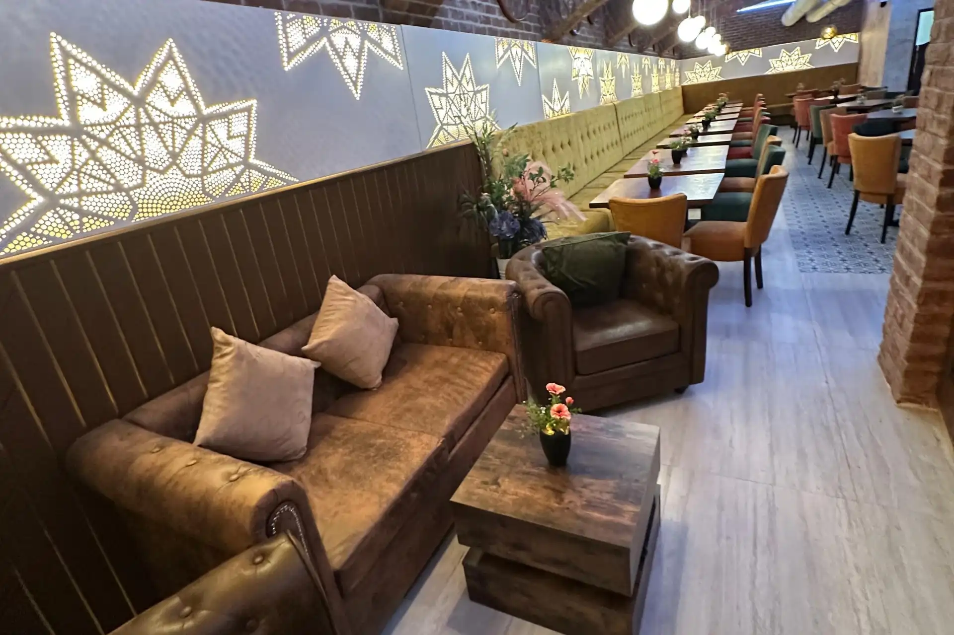 Eine gemütliche Lounge mit braunen Ledersofas, einem Couchtisch aus Holz, Reihen bunter Stühle und Tische, dekorativen Sternmustern an einer hinterleuchteten Wand und Pflanzen, die zur einladenden Atmosphäre beitragen.