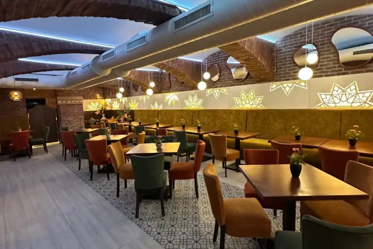 Al Zaytouna Ein gemütliches Restaurant mit Ziegelgewölbedecken, warmer Beleuchtung und geometrischen Wandbildern. Tische und Stühle in verschiedenen Farben sind ordentlich angeordnet und schaffen so einen einladenden Speiseraum mit modernem und zugleich rustikalem Flair.
