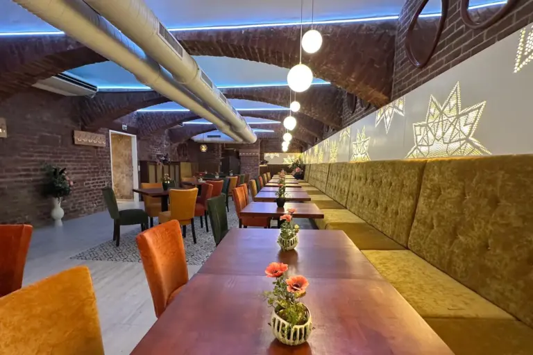 Ein gemütliches Restaurant mit langen Holztischen, bunten Stühlen, einer gewölbten Ziegeldecke, goldfarbenen gepolsterten Sitzgelegenheiten, hängenden Kugellampen und kleinen Blumenvasen auf den Tischen. Auf der rechten Seite befindet sich eine geometrische Wanddekoration.