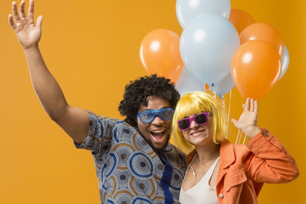 Zwei Menschen in bunten Outfits und Perücken, die lächelnd und aufgeregt mit erhobenen Armen vor orangefarbenen und blauen Luftballons posieren, bereit für Partyspiele vor einem leuchtend orangefarbenen Hintergrund.