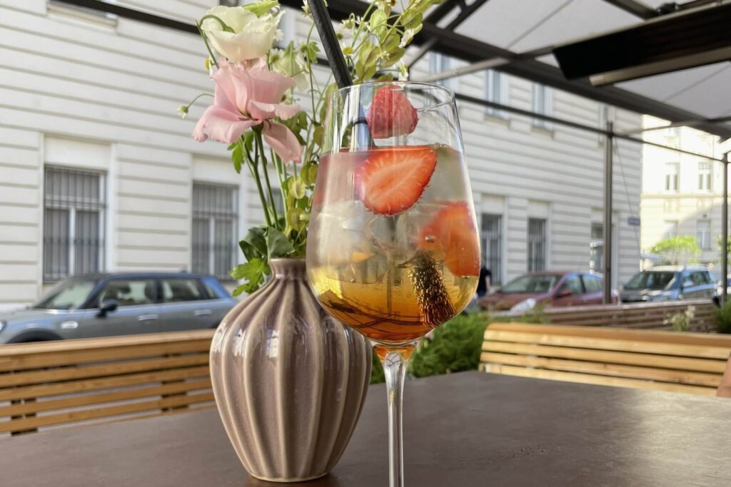 Ein Glas eisgekühlter Cocktail mit aufgeschnittenen Erdbeeren steht auf einem Schanigarten-Tisch im Freien neben einer Vase mit rosa und weißen Blumen, während im Hintergrund parkende Autos und ein Gebäude zu sehen sind.