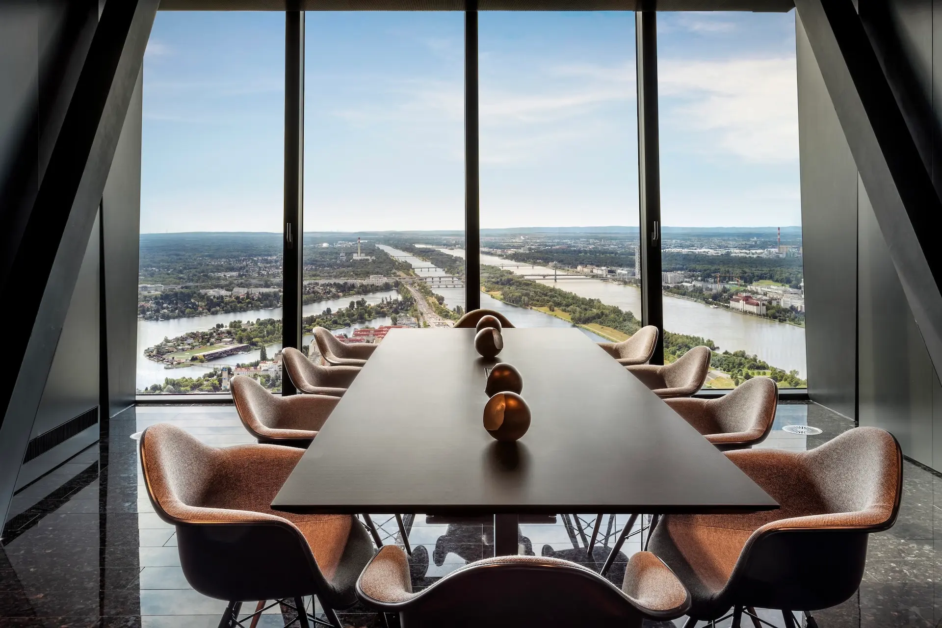 Ein moderner Konferenzraum mit einem langen Tisch, braunen Stühlen und dekorativen Kugeln verfügt über raumhohe Fenster, die an klaren Tagen einen Panoramablick auf den Fluss und die Stadtlandschaft bieten.