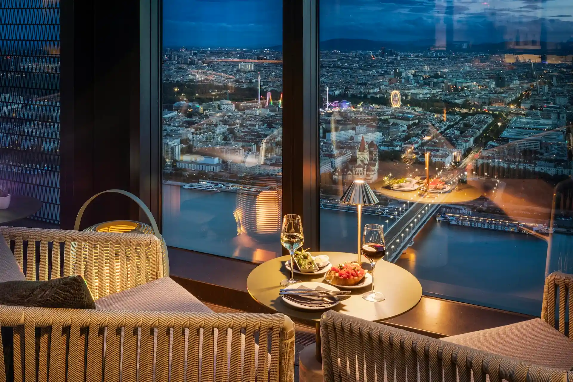 Ein gemütlicher Tisch für zwei Personen mit Wein und Vorspeisen sitzt an einem großen Fenster mit Blick auf die Skyline der Stadt und den Fluss in der Abenddämmerung, wobei sich die Lichter der Stadt auf dem Wasser spiegeln.