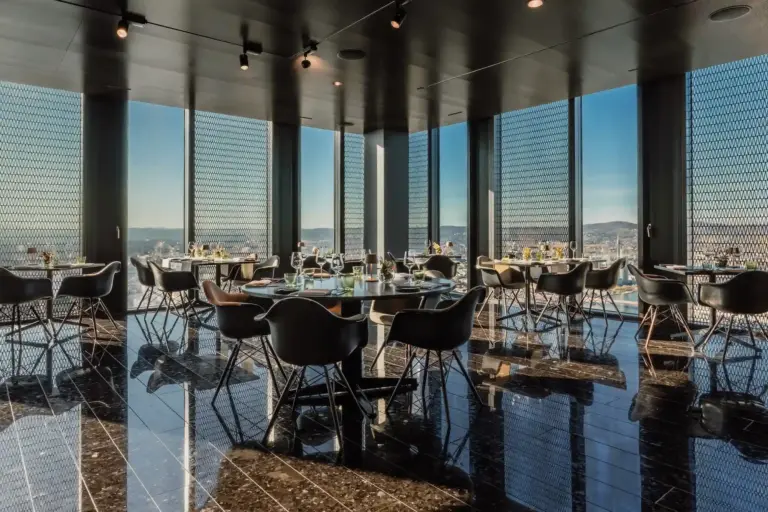 Ein modernes, gehobenes Restaurant mit schwarzen Böden, eleganten Tischen und Stühlen. Vom Boden bis zur Decke reichende Fenster bieten einen Panoramablick auf die Stadt und lassen viel natürliches Licht herein. Die Tische sind gedeckt und für die Gäste bereit.