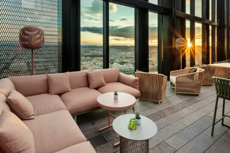 Die Lounge auf dem Dach des Altia by Urrechu verfügt über moderne rosa und beigefarbene Sitzmöbel, kleine runde Tische und raumhohe Fenster mit Blick auf die Stadt bei Sonnenuntergang. Das Sonnenlicht fällt ein und schafft eine warme und einladende Atmosphäre.
