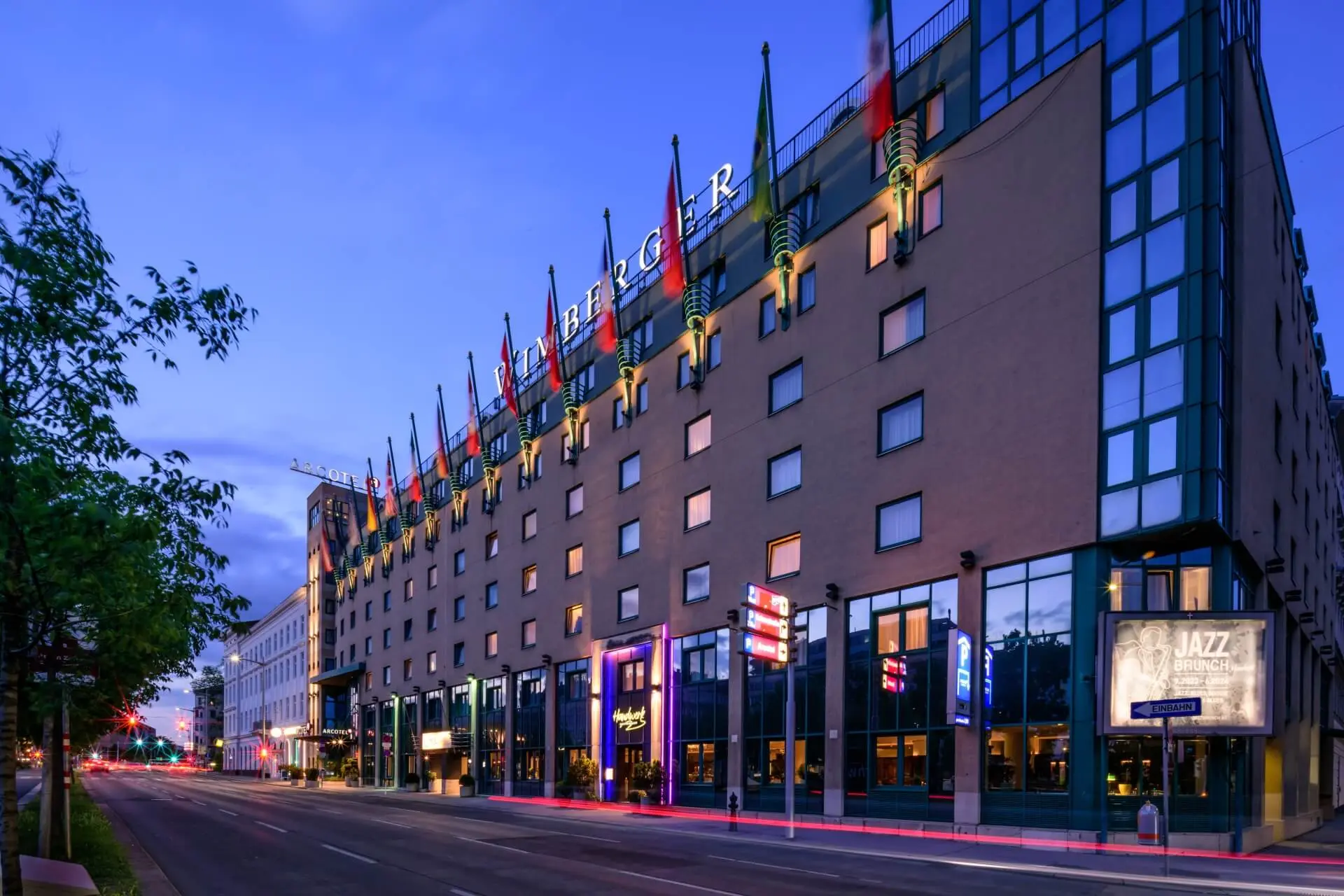 Ein modernes Hotelgebäude mit zahlreichen Nationalflaggen auf dem Dach, großen Fenstern und beleuchteten Schildern, gesehen von einer ruhigen Straße in der Abenddämmerung. Der Himmel ist klar und blau.