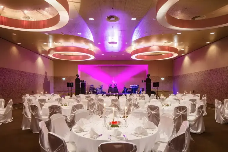 Ein für eine Veranstaltung gedeckter Bankettsaal mit runden Tischen mit weißer Bettwäsche und Stühlen, eleganten Gedecken, Blumenschmuck und einer Bühne im vorderen Bereich mit violetter Beleuchtung.