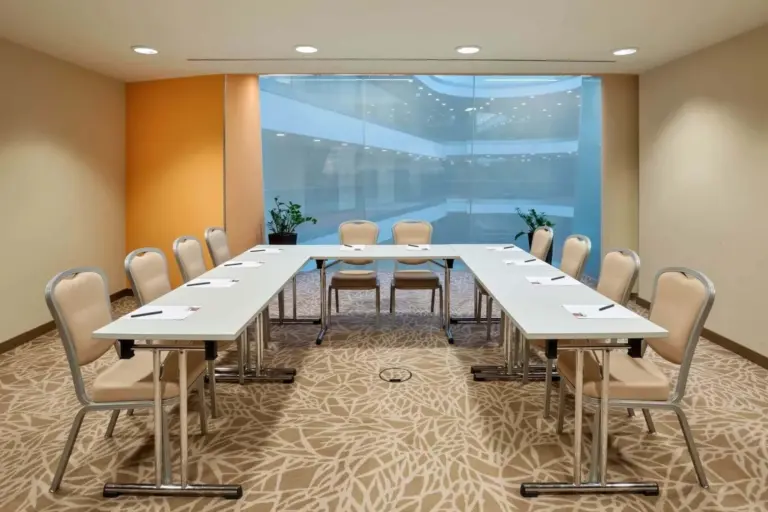 Ein moderner Konferenzraum mit beigen Stühlen, die U-förmig um drei weiße Tische angeordnet sind, vor einem großen Glasfenster und einer orangefarbenen Akzentwand. Auf jedem Tisch liegen Notizblöcke und Stifte bereit.