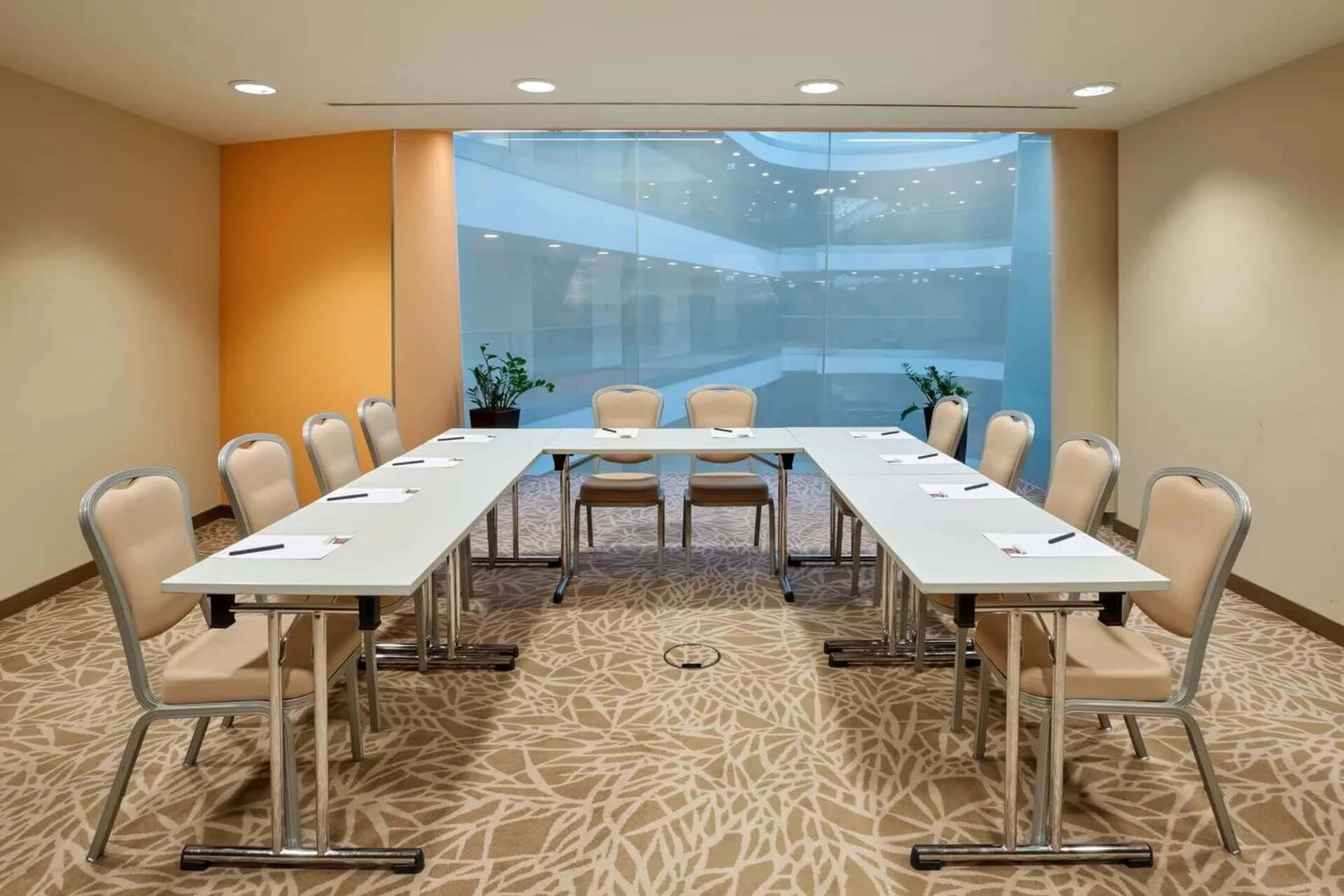 Ein moderner Konferenzraum mit beigen Stühlen, die U-förmig um drei weiße Tische angeordnet sind, vor einem großen Glasfenster und einer orangefarbenen Akzentwand. Auf jedem Tisch liegen Notizblöcke und Stifte bereit.
