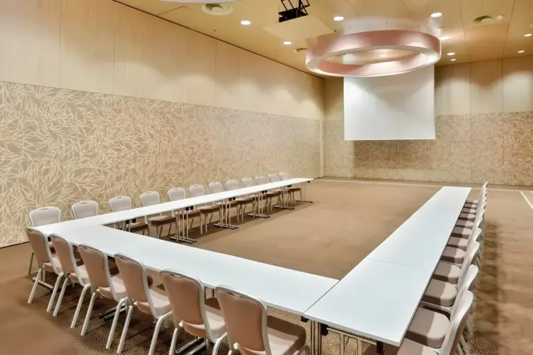 Ein geräumiger Konferenzraum mit U-förmigen weißen Tischen, beigen Stühlen, gemusterten Wänden, einem Deckenprojektor und einer großen Projektionsfläche an der Stirnseite. Der Raum wirkt sauber und gut beleuchtet.