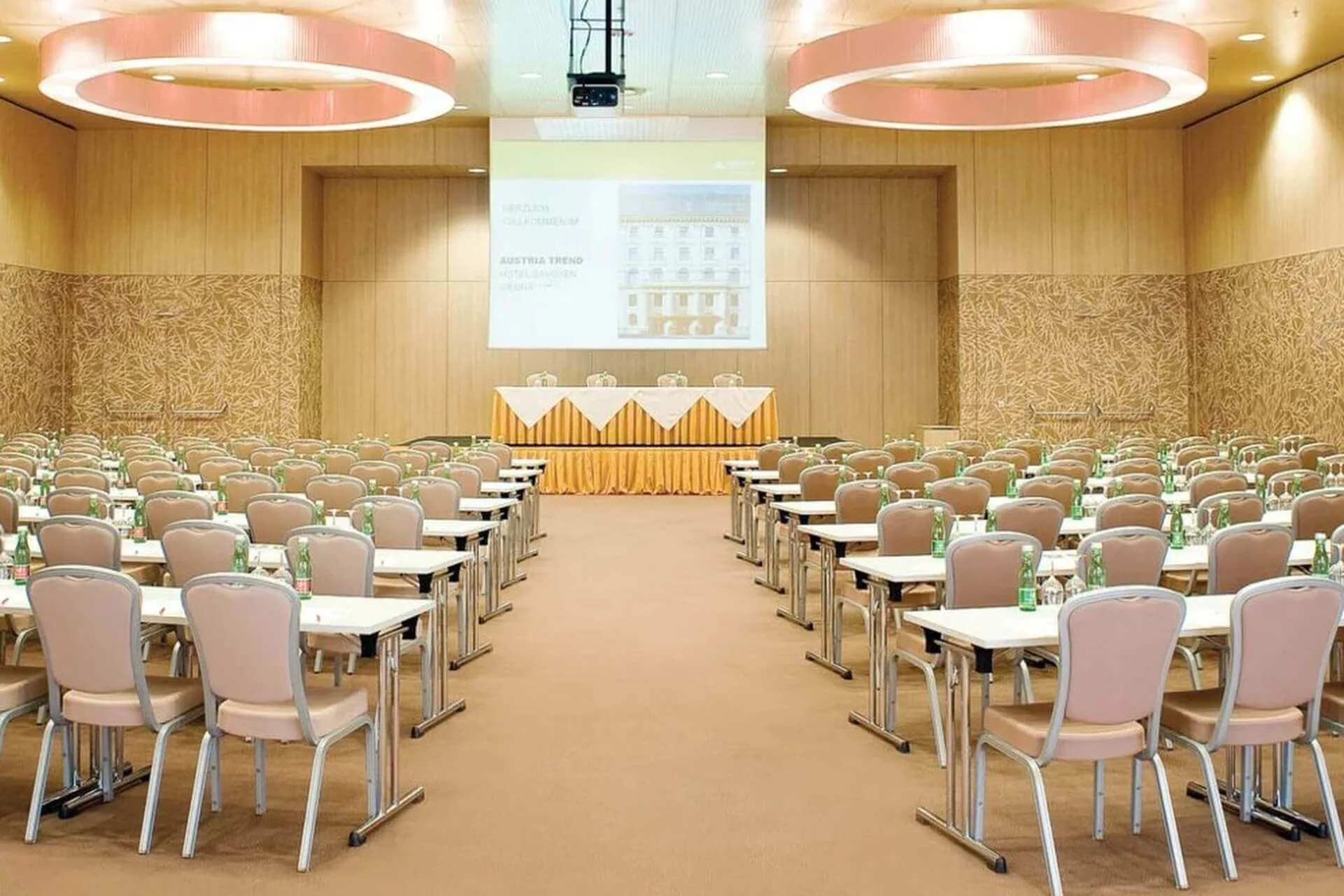 Ein geräumiger Konferenzraum mit Reihen beiger Stühle und Tische vor einer Bühne mit drei Stühlen und einem Tisch mit gelben Röcken. An der Stirnseite ist eine große Projektionsfläche angebracht. Auf jedem Tisch stehen Wasserflaschen.