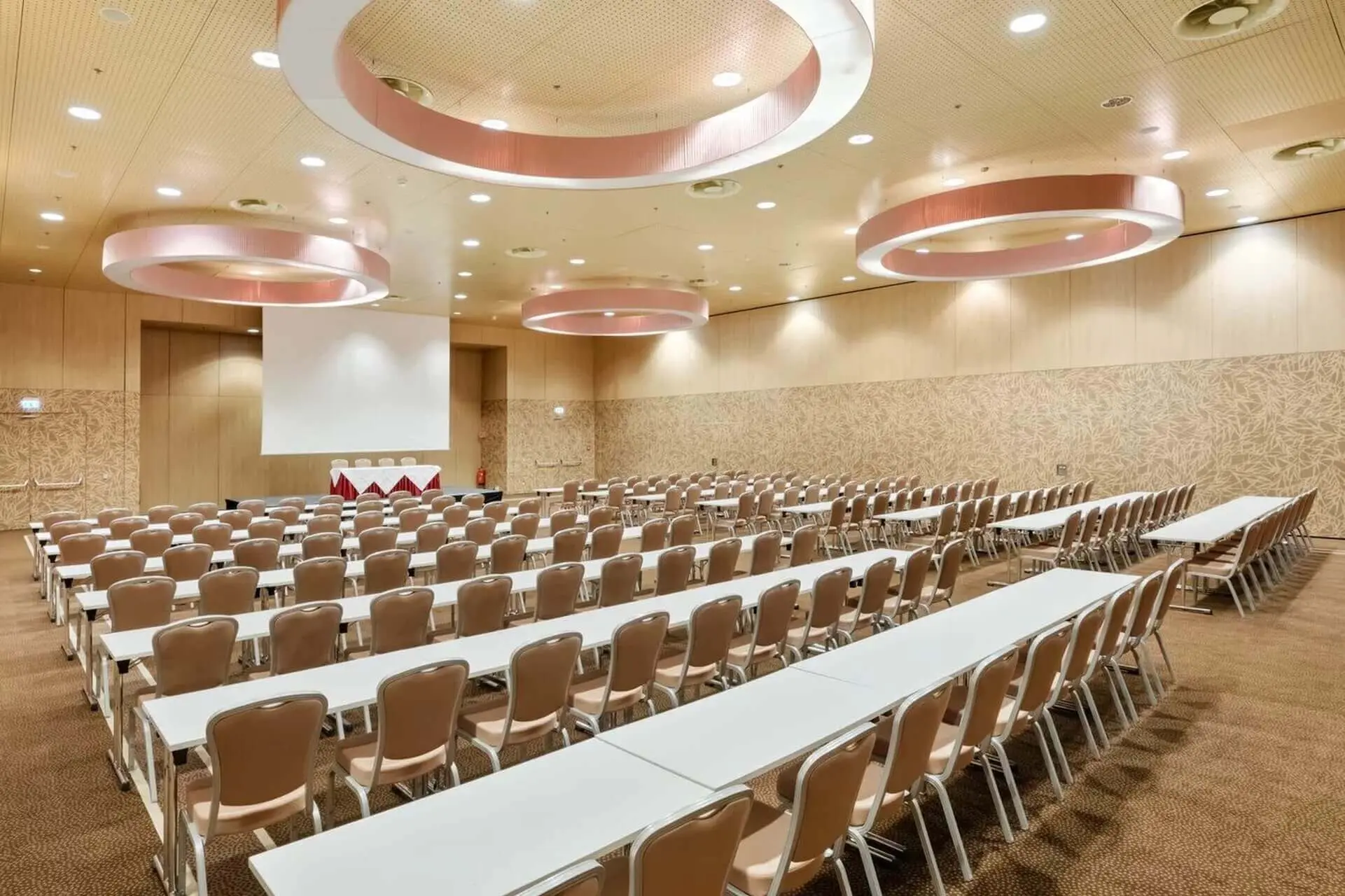 Ein großer, moderner Konferenzraum mit Reihen von weißen Tischen und beigen Stühlen vor einer Projektionsfläche. Darüber hängen runde Deckenleuchten, und die Wände sind mit einem hellen Muster versehen.