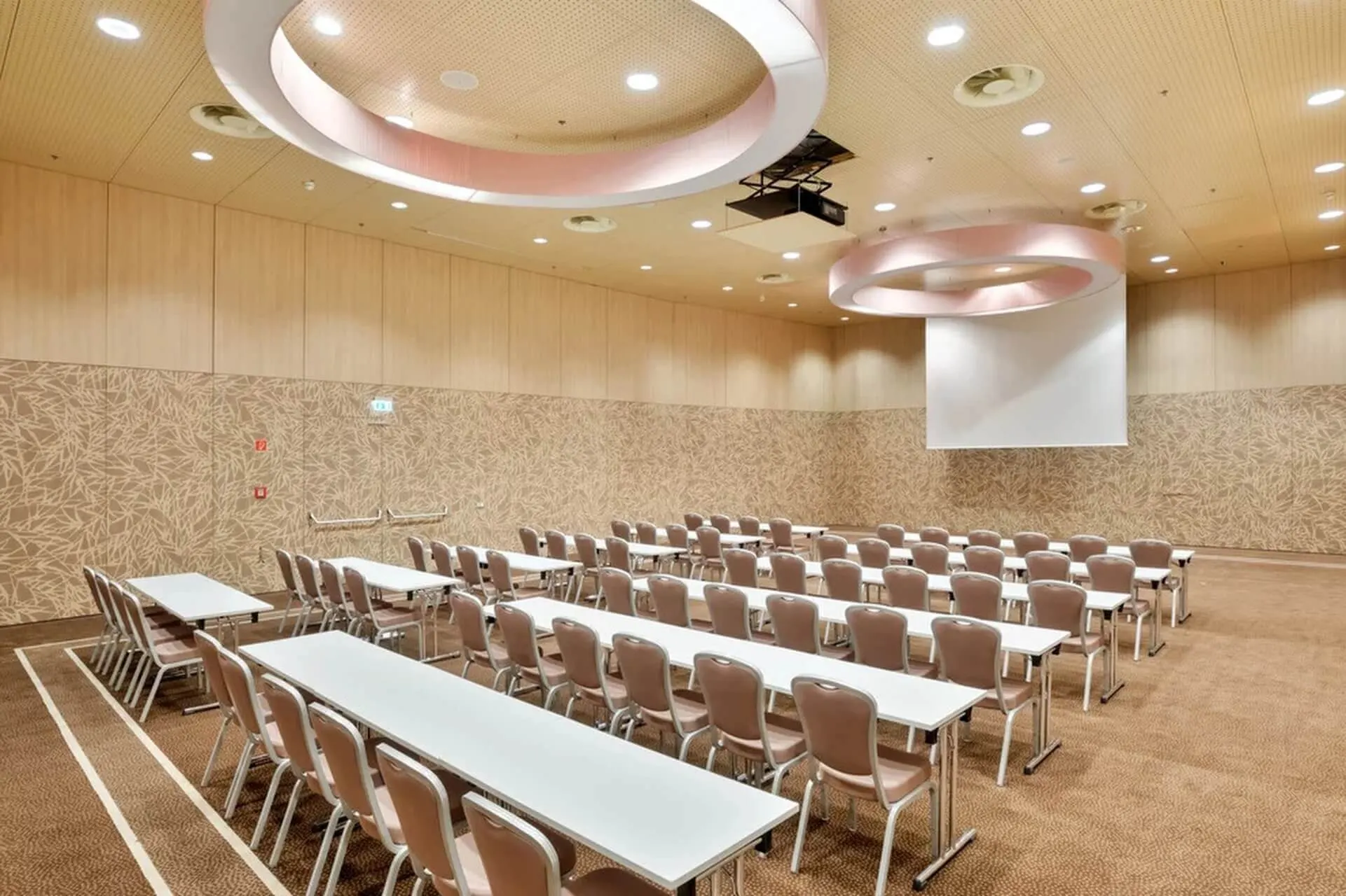 Ein geräumiger Konferenzraum mit Reihen von weißen Tischen und beigen Stühlen vor einer großen Projektionsfläche. Der Raum hat gemusterte Wände, Teppichböden und moderne runde Deckenleuchten.
