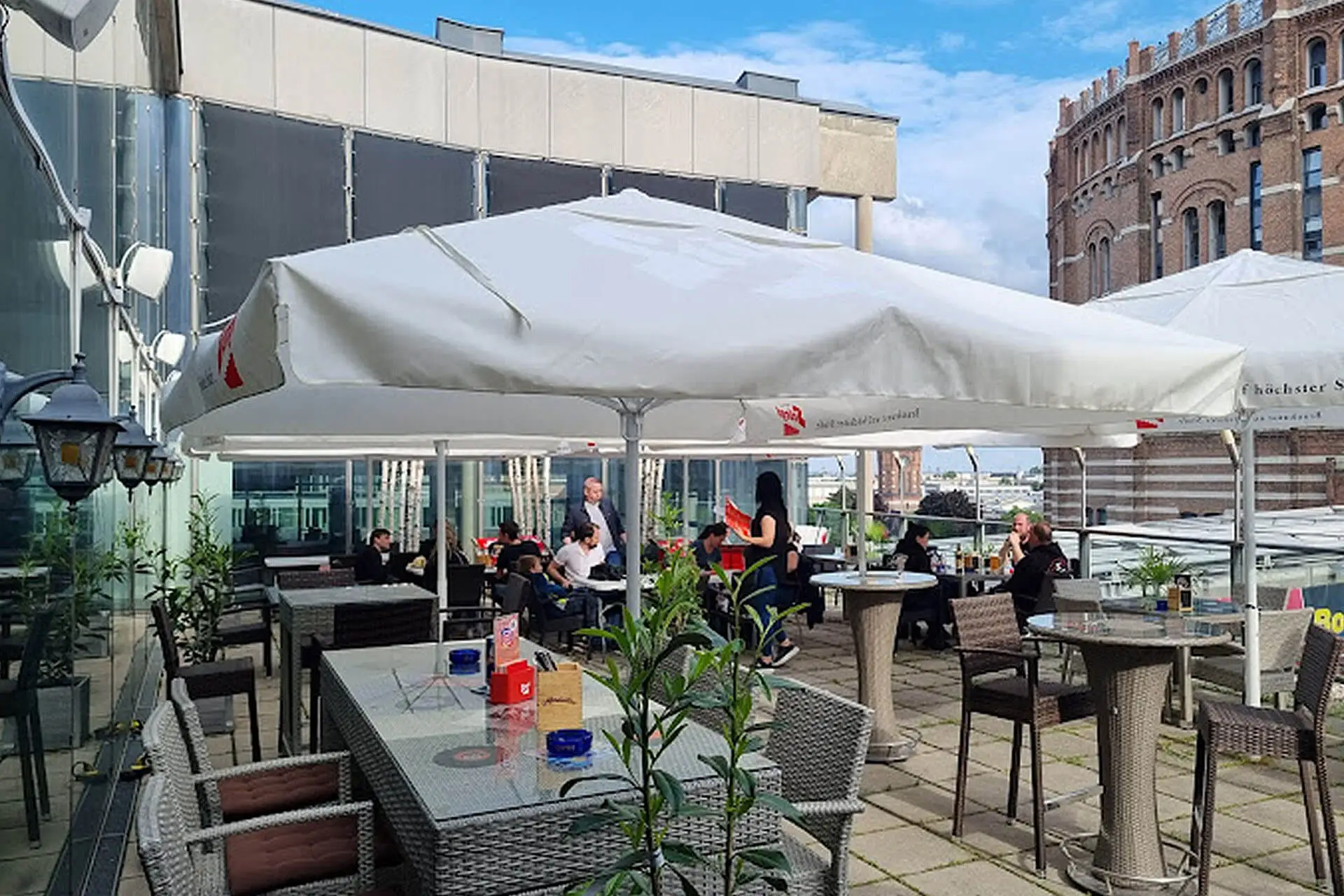 Menschen sitzen an Tischen auf einer modernen Außenterrasse mit großen weißen Sonnenschirmen, umgeben von Glasgebäuden und einem historischen Backsteinbau im Hintergrund, unter einem blauen Himmel mit einigen Wolken.