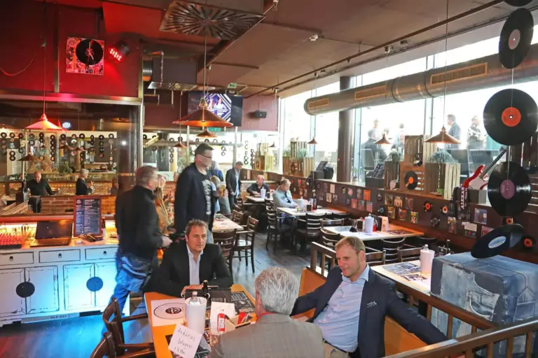 Ein lebhaftes Restaurant mit einem Retro-Musik-Thema, mit Vinyl-Schallplatten an den Wänden. Mehrere Leute sitzen an Tischen, während andere stehen und sich unterhalten. Große Fenster lassen natürliches Licht herein. Drei Männer in Anzügen unterhalten sich an einem der vorderen Tische.