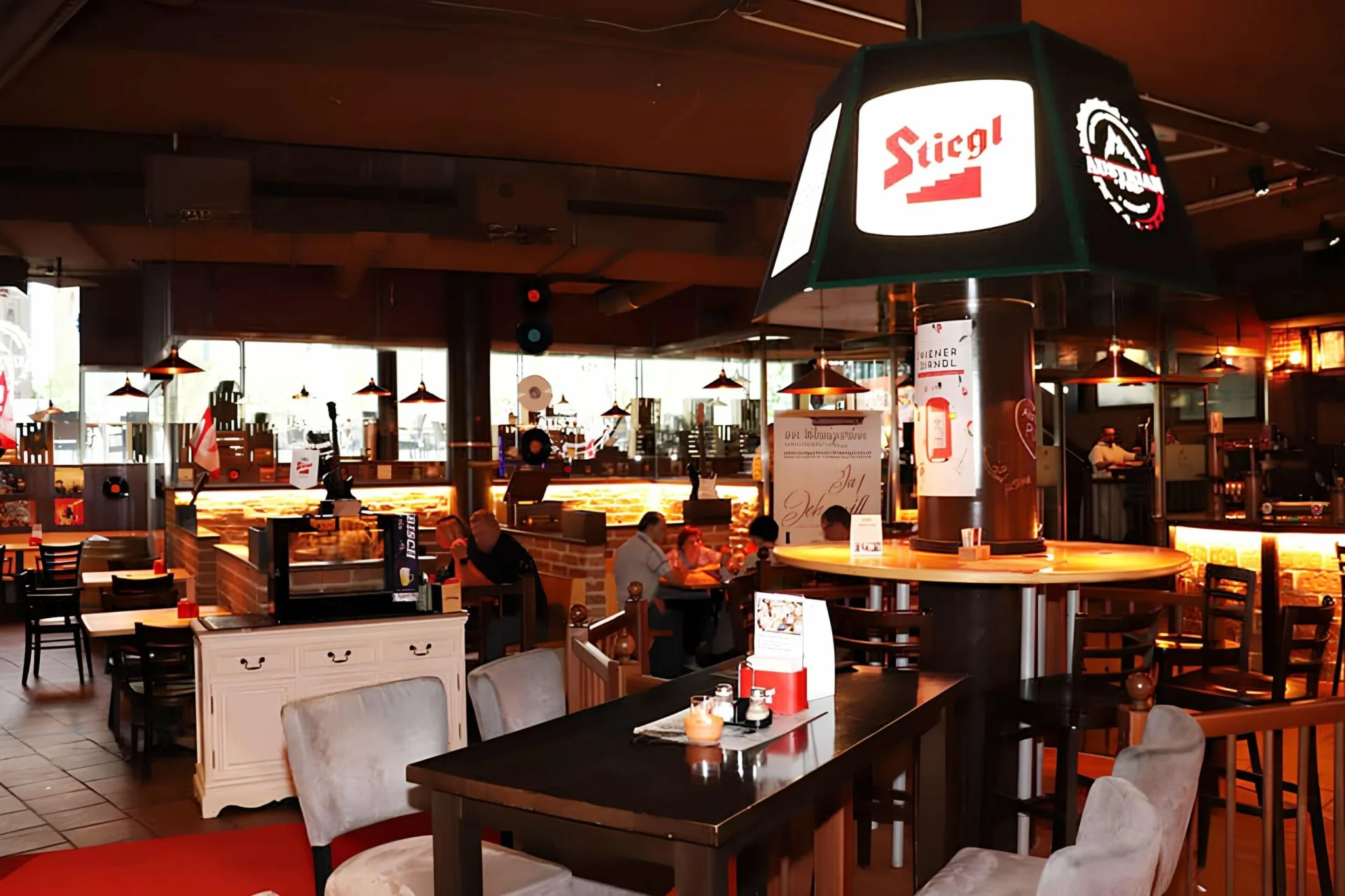 Ein gemütliches Restaurant mit Holztischen und -stühlen, warmer Beleuchtung und einer großen Hängelampe mit einem Stiegl-Logo über einem Tisch. Im Hintergrund sitzen mehrere Personen beim Essen.