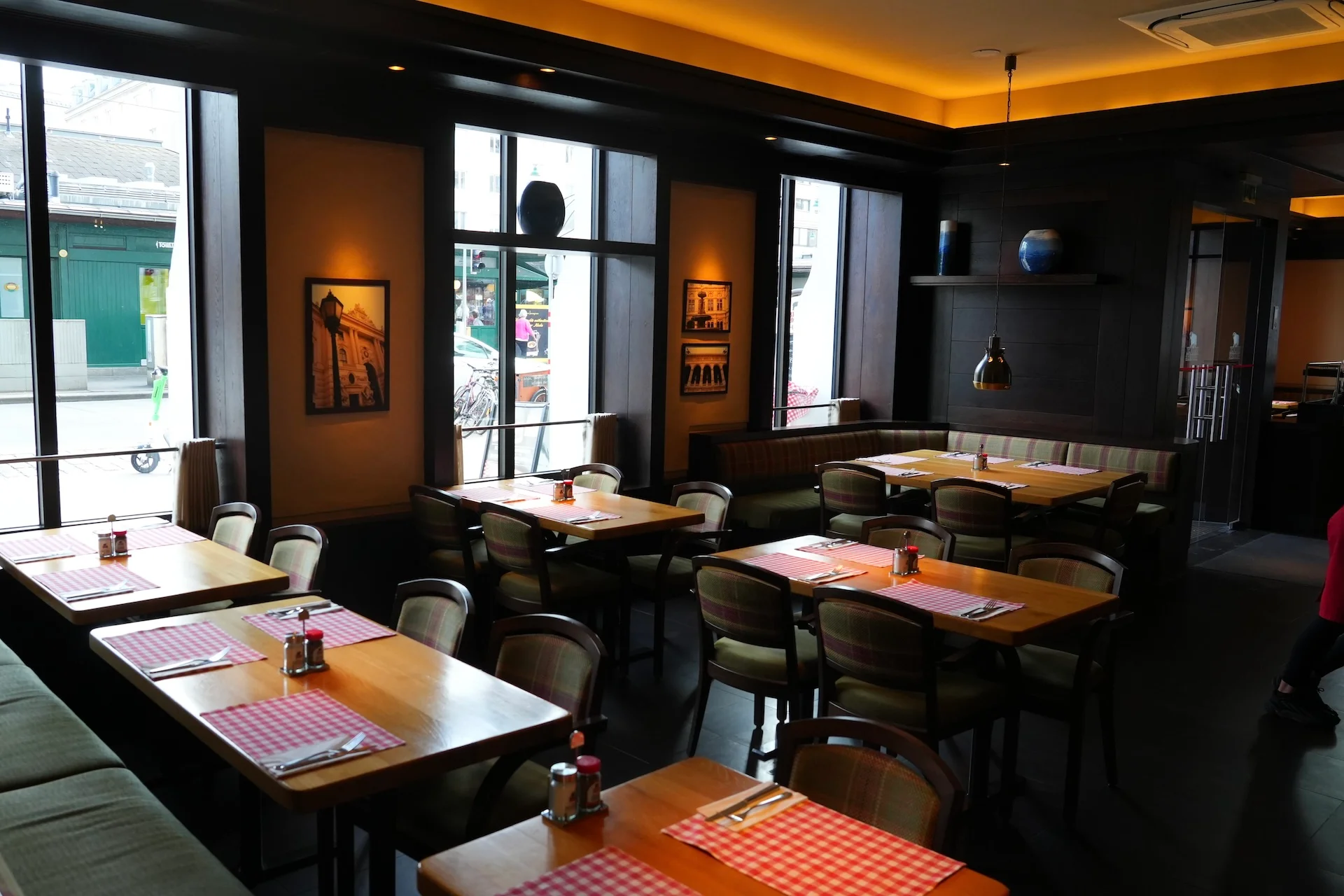 Ein gemütliches Restaurant mit Holztischen und -stühlen, rot-karierten Tischsets, Gewürzflaschen und großen Fenstern, die den Raum mit natürlichem Licht durchfluten. Gerahmte Kunstwerke schmücken die Wände.