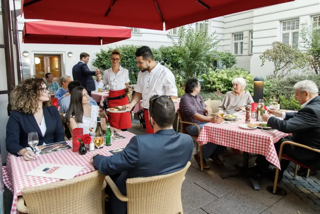 Eine Gruppe von Menschen speist an Tischen im Freien mit rot karierten Tischdecken; Kellner servieren Speisen und Getränke unter roten Sonnenschirmen in einem begrünten Innenhof mit umliegenden Gebäuden.