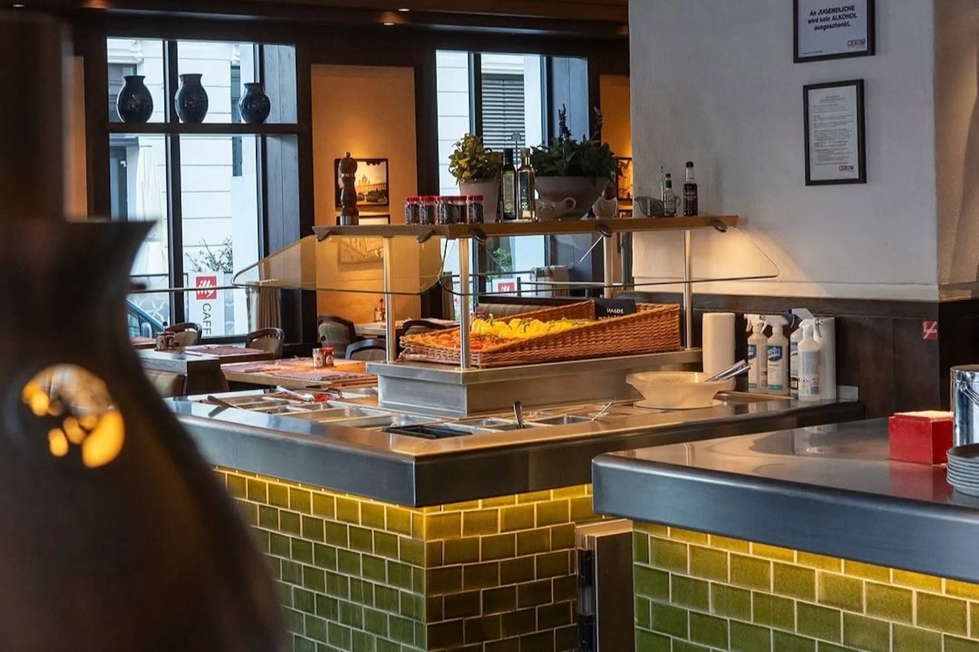 Ein hell erleuchtetes Restaurantbuffet mit einem Tresen mit grünen Fliesen, Tabletts mit Speisen unter Wärmelampen und verschiedenen Gewürzen und Reinigungsmitteln auf dem Tresen. Im Hintergrund sind große Fenster und Dekoration zu sehen.