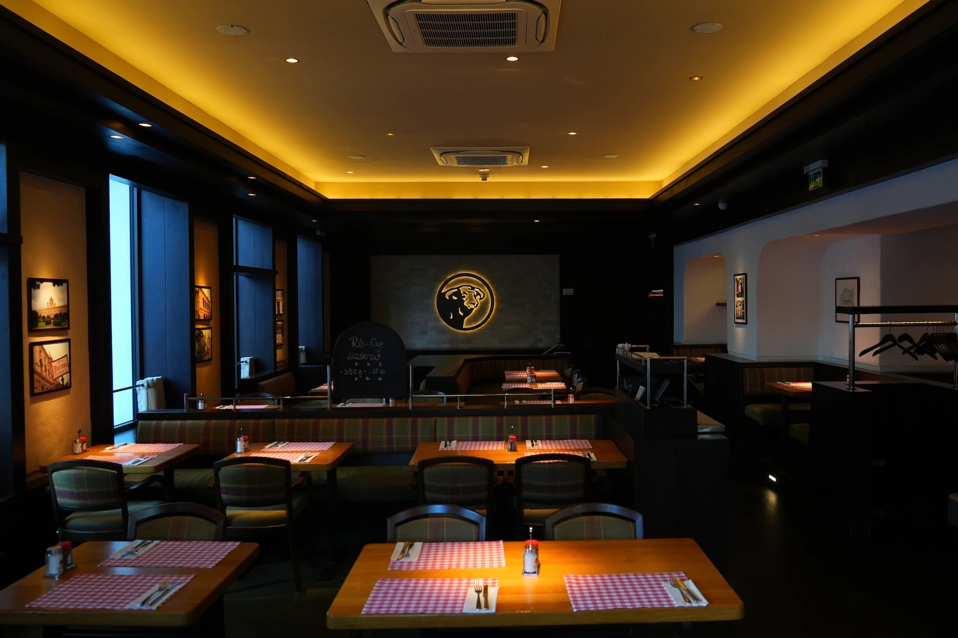 Ein gemütliches, schwach beleuchtetes Restaurant mit Holztischen, die mit rot-karierten Tischsets gedeckt sind, gepolsterten Stühlen und einem leuchtenden Logo an der Rückwand, das eine warme und einladende Atmosphäre schafft.