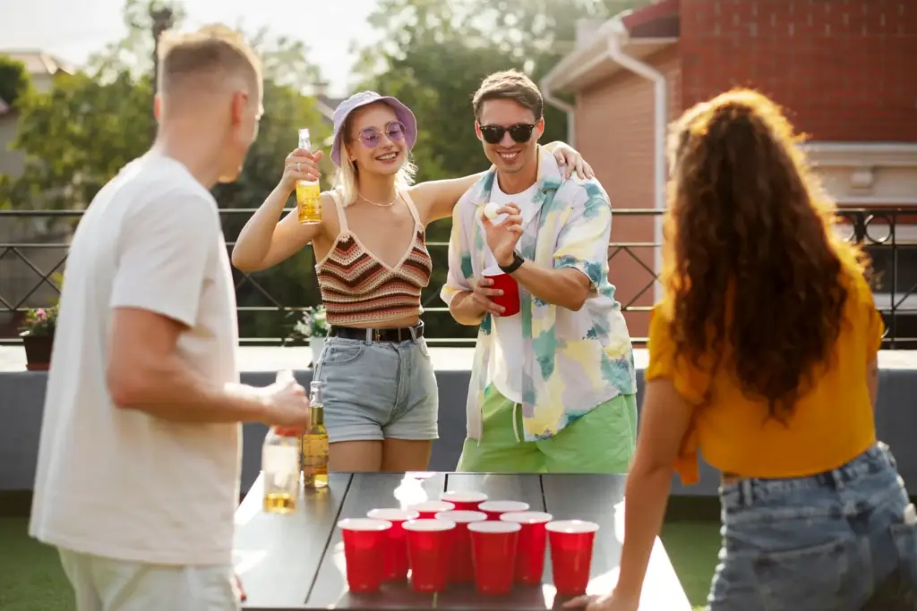 Vier junge Erwachsene genießen die Partyspiele für Erwachsene und spielen an einem sonnigen Tag im Freien Bierpong. Sie lächeln, halten Getränke in der Hand, und auf dem Tisch vor ihnen stehen rote Becher.