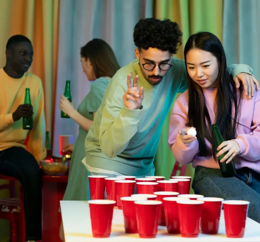 Eine Gruppe junger Erwachsener vergnügt sich bei Partyspielen für Erwachsene und spielt Bierpong. Zwei im Vordergrund zielen mit einem Tischtennisball zwischen roten Bechern, während andere im Hintergrund Getränke halten und sich unterhalten.
