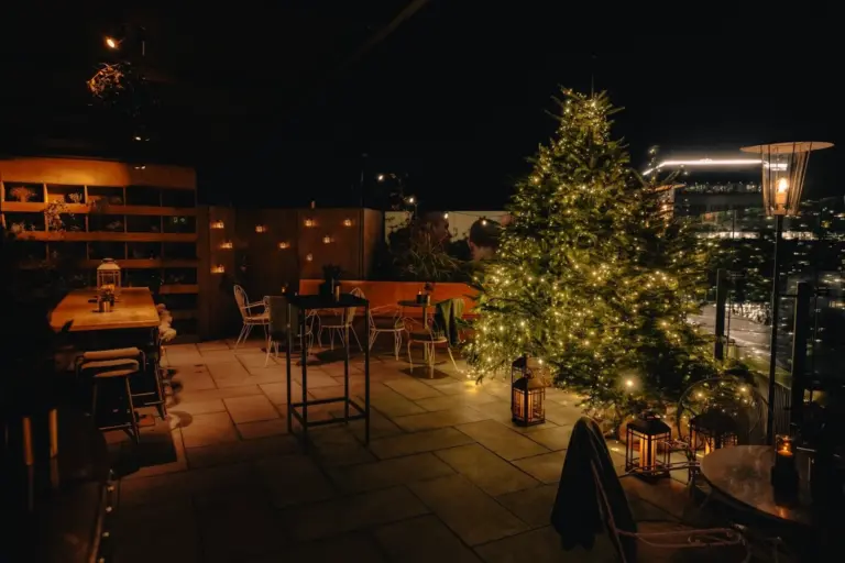 Eine Dachterrasse ist nachts mit einem beleuchteten Weihnachtsbaum, Lichterketten, Holztischen, Stühlen und Laternen geschmückt, was eine warme, festliche Atmosphäre schafft.