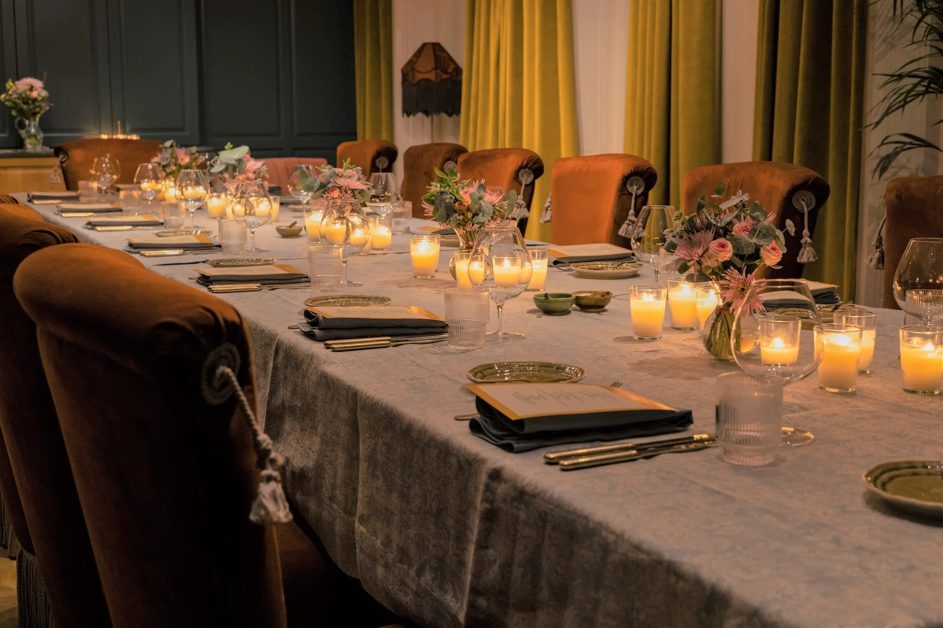 Ein langer Esstisch mit Kerzen, Blumengestecken, eleganten Gedecken und braunen Samtstühlen in einem sanft beleuchteten, edlen Raum mit gelben Vorhängen.
