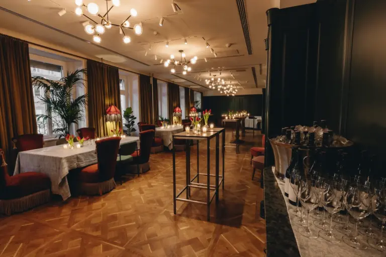 Ein eleganter Speisesaal mit Holzböden, mit weißen Tischdecken gedeckten Tischen, roten Stühlen, Blumengestecken, modernen Kronleuchtern und einem Barbereich, an dessen Seite Weinflaschen und Gläser stehen. Warme, stimmungsvolle Beleuchtung erfüllt den Raum.