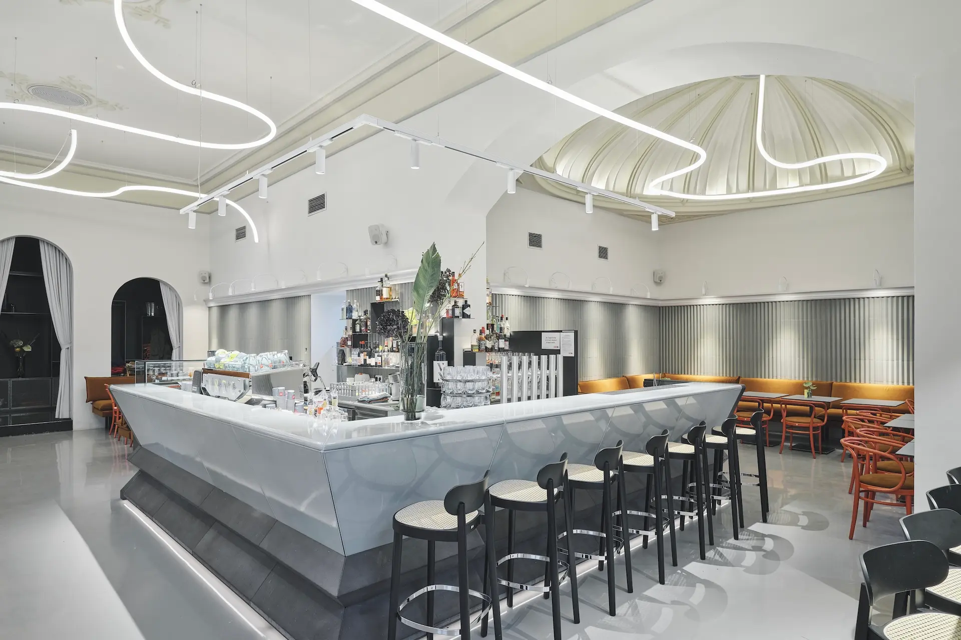 Eine moderne, hell erleuchtete Bar mit einem geometrischen weißen Tresen, schwarzen Barhockern, orangefarbenen Stühlen und geschwungenen Neon-Deckenleuchten. Hohe Decken und drapierte Stoffe verleihen der modernen Einrichtung einen eleganten Touch.