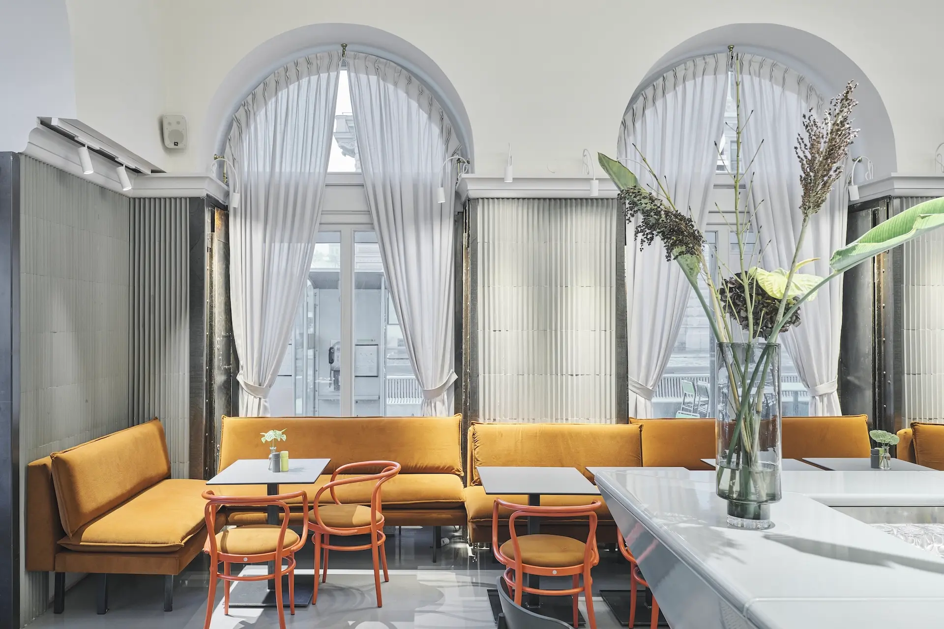 Ein modernes Café-Interieur mit senfgelben Sofas, orangefarbenen Stühlen, kleinen weißen Tischen, hohen Bogenfenstern mit durchsichtigen Vorhängen und einer Vase mit Blumen auf einem Tresen im Vordergrund.