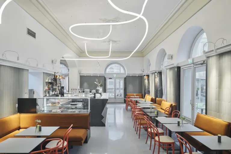 Modernes Café-Interieur mit hohen Decken, geschwungenen LED-Leuchten, orangefarbenen Sitzpolstern, roten Stühlen und großen Bogenfenstern, die natürliches Licht hereinlassen. Auf der linken Seite befinden sich eine Vitrine und eine Kaffeebar.
