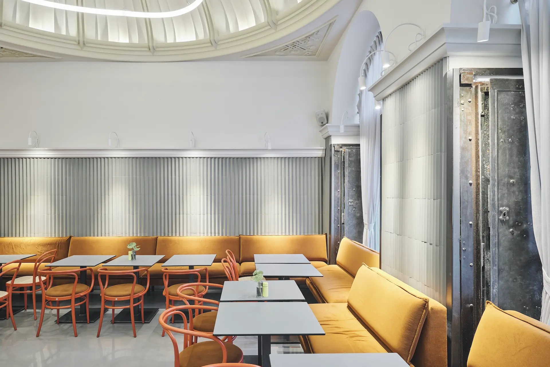 Ein modernes Café mit senfgelben Sitzpolstern, orangefarbenen Stühlen, weißen Tischen und einer geschwungenen Glasdecke. Sanfte Beleuchtung und minimale Dekoration schaffen eine helle, einladende Atmosphäre. Kleine Pflanzen stehen auf den Tischen.