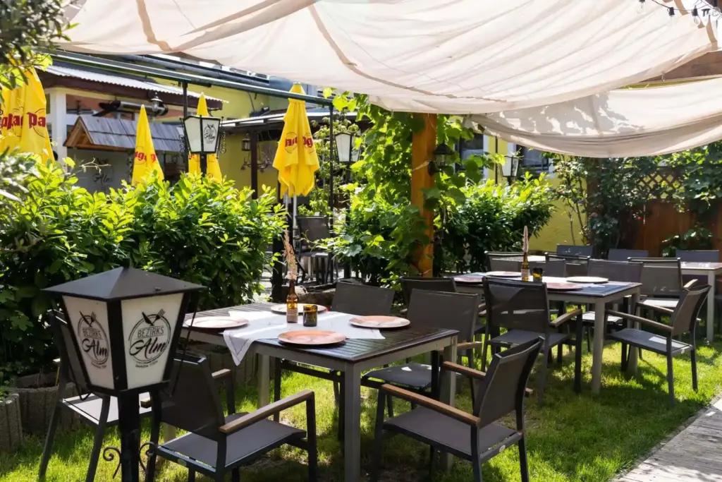 Eine Restaurantterrasse im Freien mit Tischen zum Essen, beschattet von einem weißen Vordach. Grünpflanzen umgeben den Bereich, und im Hintergrund sind gelbe Sonnenschirme zu sehen, die eine gemütliche, einladende Atmosphäre schaffen.