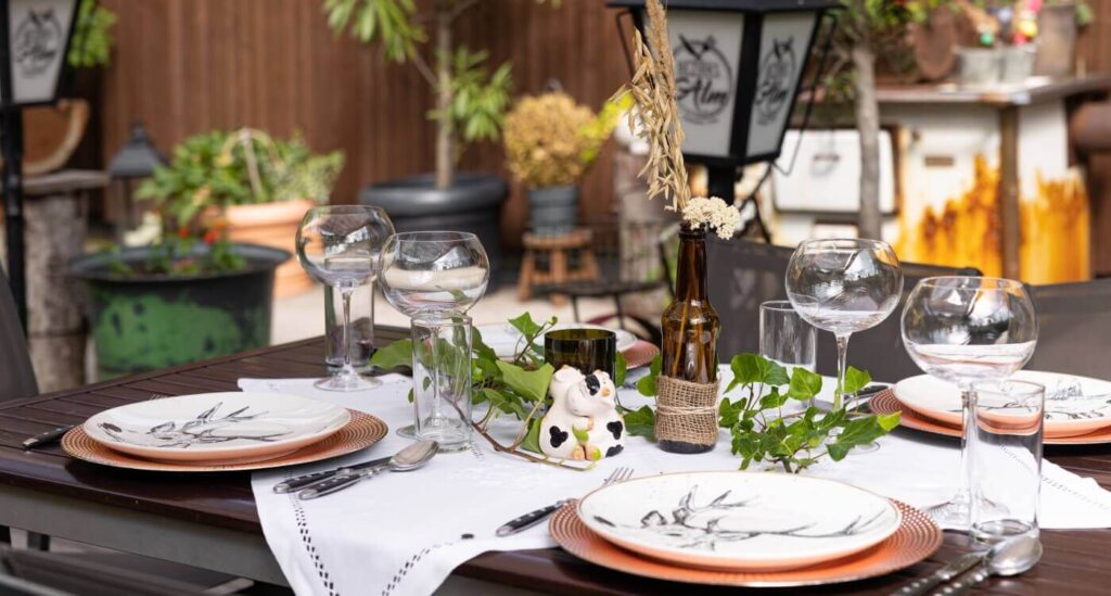 Ein ordentlich gedeckter Esstisch im Freien mit Tellern, Weingläsern, Besteck, einer Tischdecke und einer kleinen Kuhfigur schafft eine einladende Szene - ideal für eine Gartenfeier oder Hochzeitslocation Wien, umgeben von Topfpflanzen und üppigem Grün.