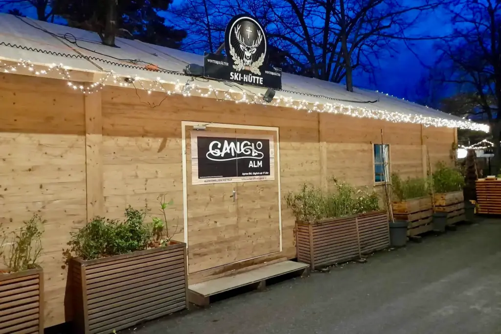 Ein Holzgebäude, das nachts mit Lichterketten geschmückt ist, mit einem Schild mit der Aufschrift GANG ALM Ski-Hütte über dem Eingang und Blumenkästen an der Vorderseite. Im Hintergrund sind blattlose Bäume zu sehen.