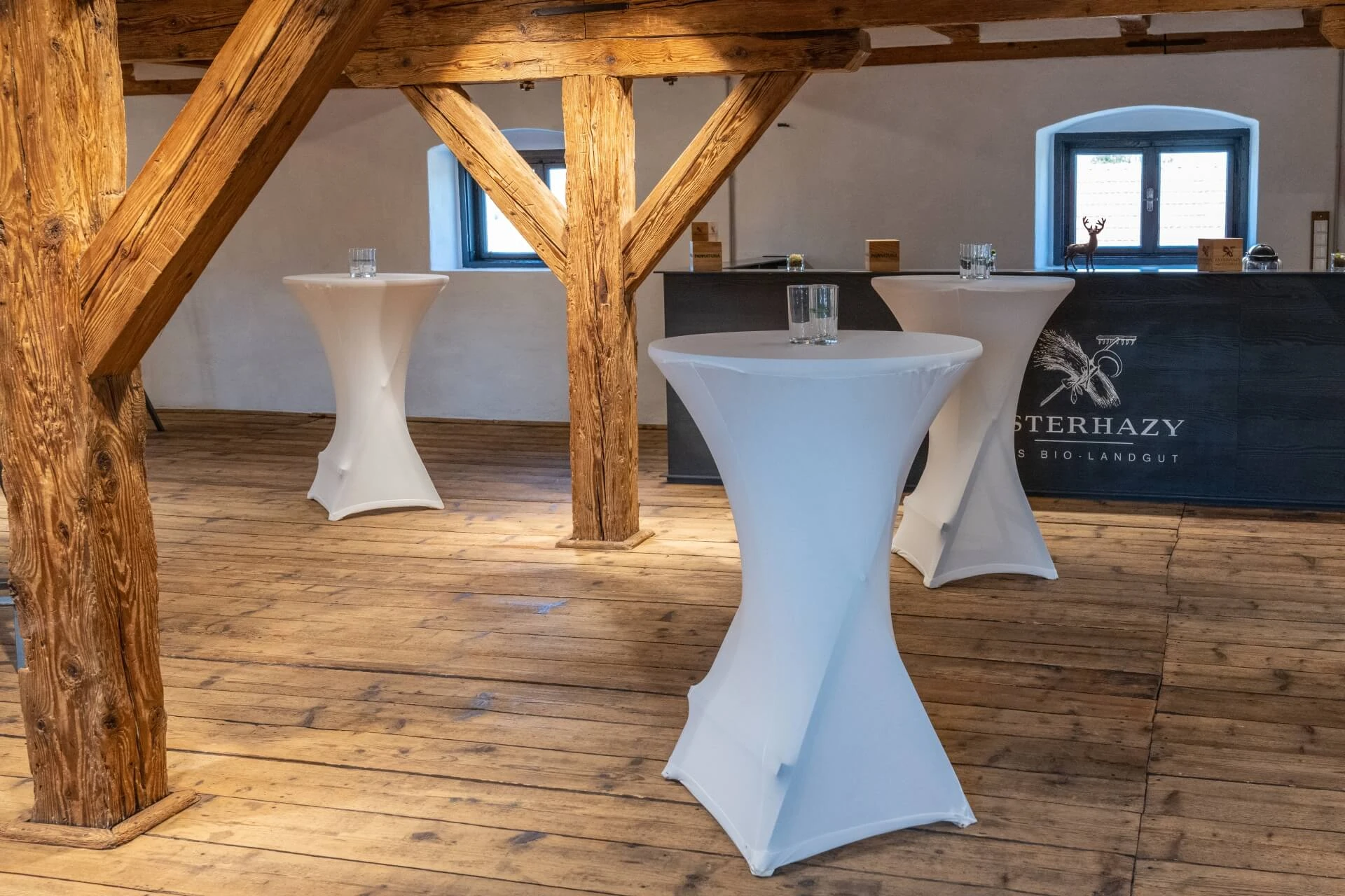 In einem rustikalen Raum mit Holzböden und -balken stehen hohe, mit weißen Tüchern bedeckte Cocktailtische, auf denen jeweils ein Glas Wasser steht, und eine schwarze Theke mit der Aufschrift ESTERHAZY BIO-LANDGUT im Hintergrund.