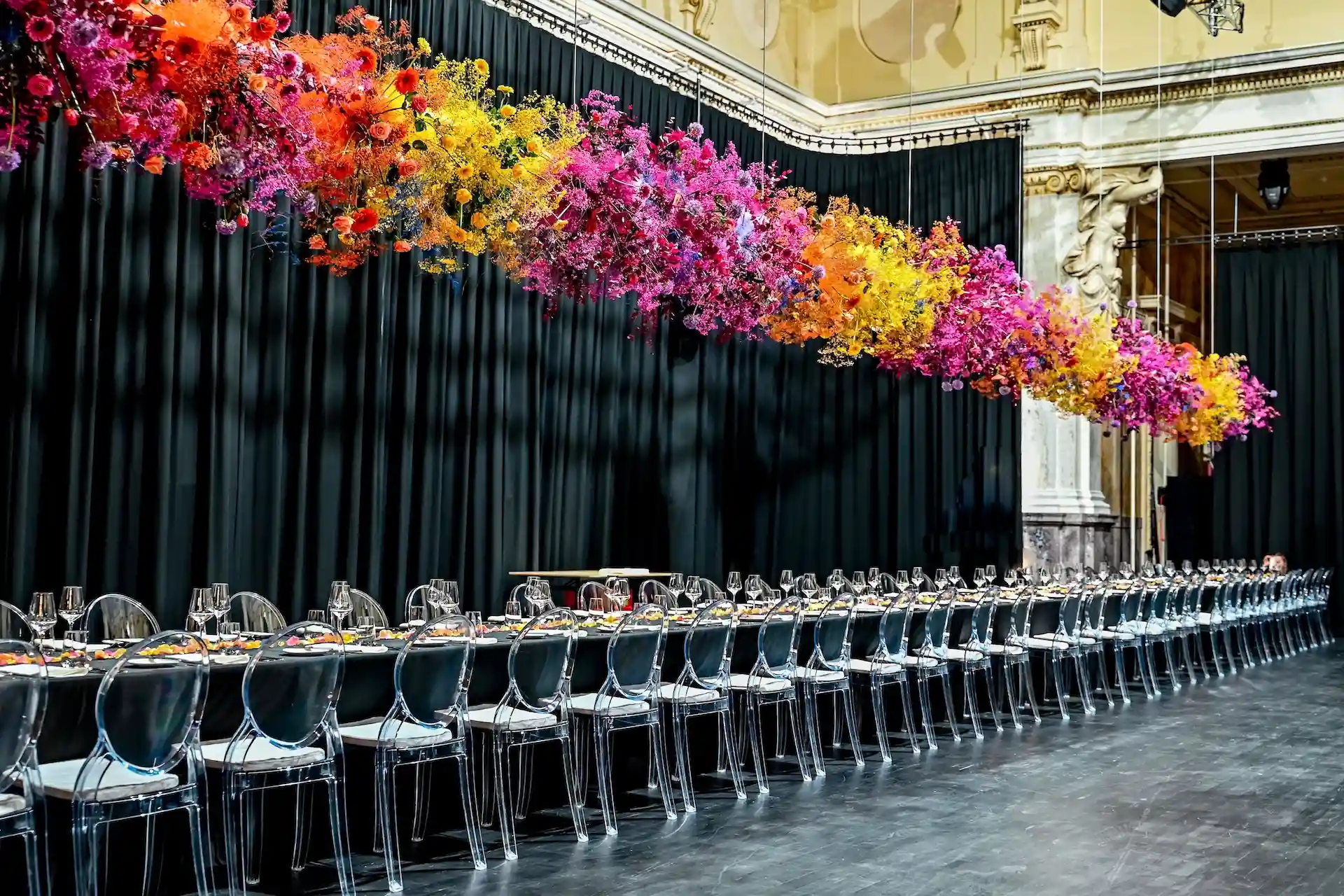 In einem großen, eleganten Raum mit hohen Decken und schwarzen Vorhängen steht ein langer Esstisch mit schwarzen Stühlen unter einem eindrucksvollen, hängenden Blumenarrangement aus bunten Blüten in Orange-, Rosa- und Gelbtönen.