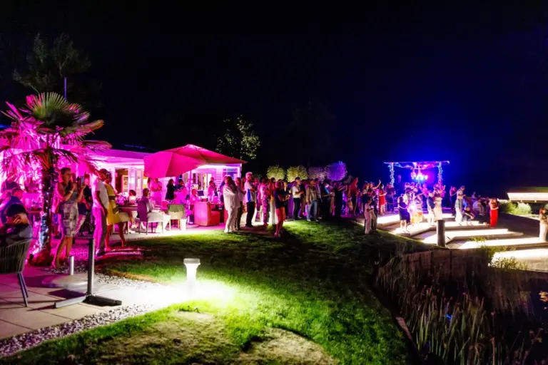 Eine lebhafte nächtliche Party im Freien mit Menschen, die sich auf einer Wiese treffen, bunten rosa und blauen Lichtern, die die Szene beleuchten, und Tischen mit Sonnenschirmen neben einem modernen Gebäude.