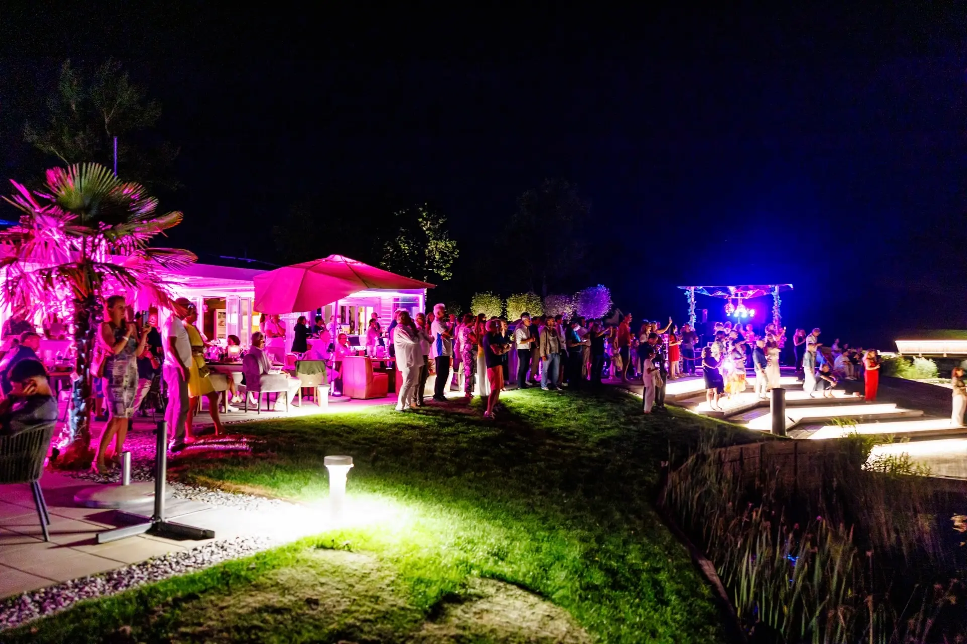 Eine lebhafte nächtliche Party im Freien mit Menschen, die sich auf einer Wiese treffen, bunten rosa und blauen Lichtern, die die Szene beleuchten, und Tischen mit Sonnenschirmen neben einem modernen Gebäude.