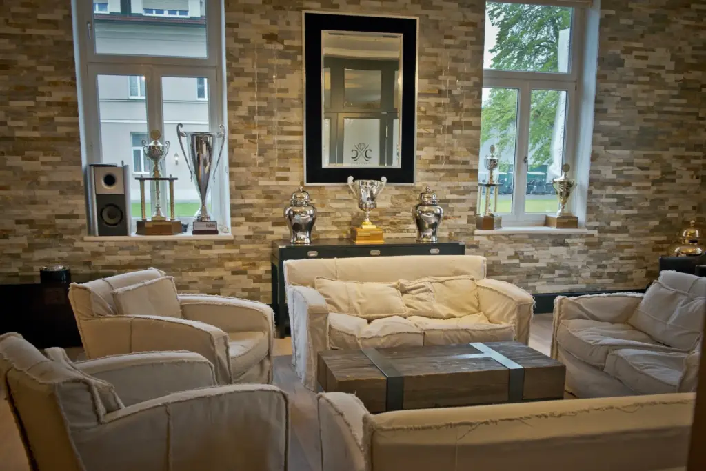 Eine gemütliche Bootshaus-Lounge mit vier beigen Sesseln und einem Sofa um einen hölzernen Couchtisch. Dahinter sind mehrere Trophäen in Regalen vor einer Backsteinwand mit großen Fenstern und einem zentralen schwarz gerahmten Spiegel ausgestellt.