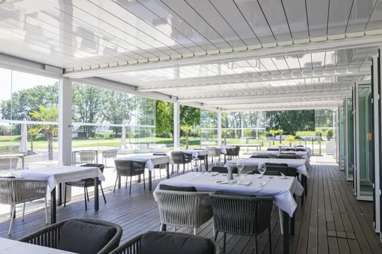 Eine Restaurantterrasse im Freien mit ordentlich gedeckten Tischen und Stühlen, überdacht von einer modernen weißen Pergola. Im Hintergrund sind große Fenster und Grünflächen zu sehen, die eine offene, luftige und einladende Atmosphäre zum Essen schaffen.
