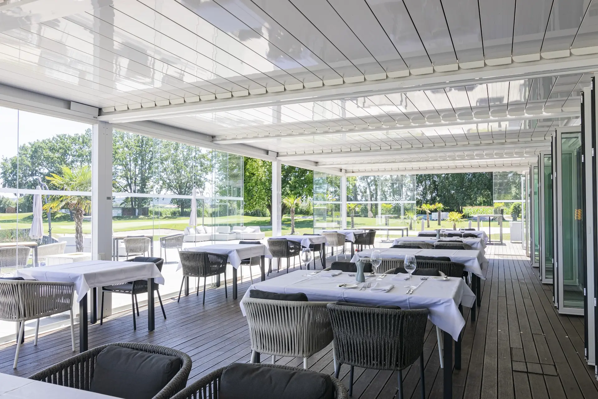 Eine Restaurantterrasse im Freien mit ordentlich gedeckten Tischen und Stühlen, überdacht von einer modernen weißen Pergola. Im Hintergrund sind große Fenster und Grünflächen zu sehen, die eine offene, luftige und einladende Atmosphäre zum Essen schaffen.