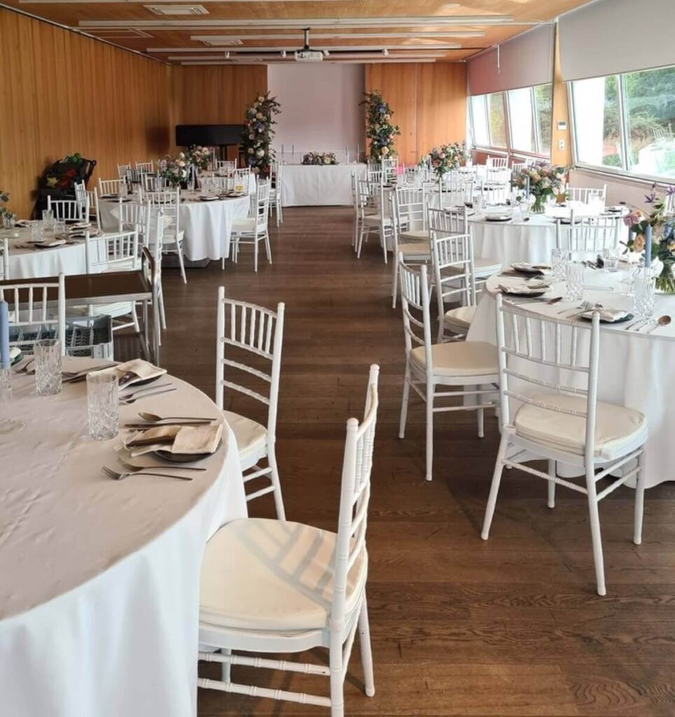 Ein heller Bankettsaal mit runden Tischen mit weißen Tischdecken, die für Veranstaltungen gedeckt sind. Weiße Stühle und florale Tafelaufsätze sorgen für Eleganz. Große Fenster säumen eine Seite und machen ihn zu einem idealen Partyraum mieten Wien für Versammlungen.