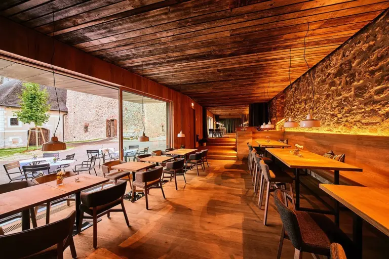Modernes Restaurant mit Holzböden, Decken und Steinwänden. Große Fenster bieten einen Blick auf einen Außenbereich mit Tischen und Stühlen. Die warme Beleuchtung schafft eine gemütliche Atmosphäre.